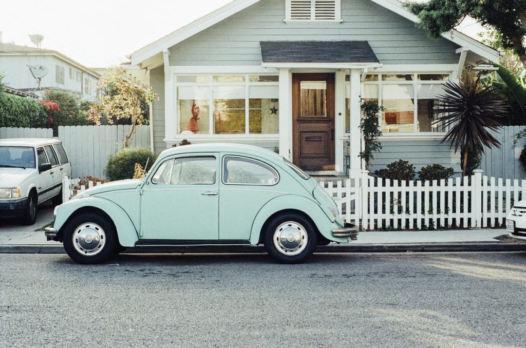 A VW Beetle in front of a single-story home with a white fence.