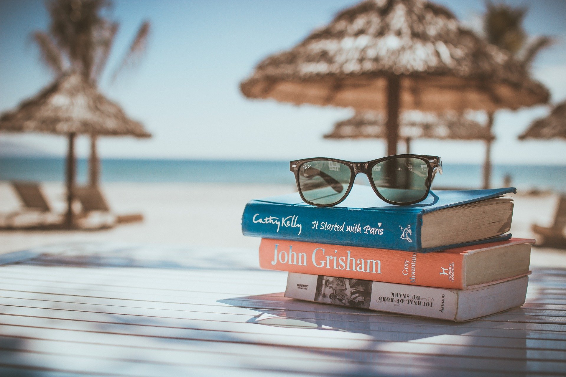 A stack of books with a pair of sunglasses on top with a background of a beach.