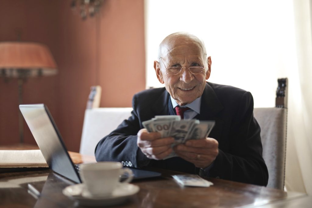 Elderly man in a suit holding money at a desk with a laptop and a teacup