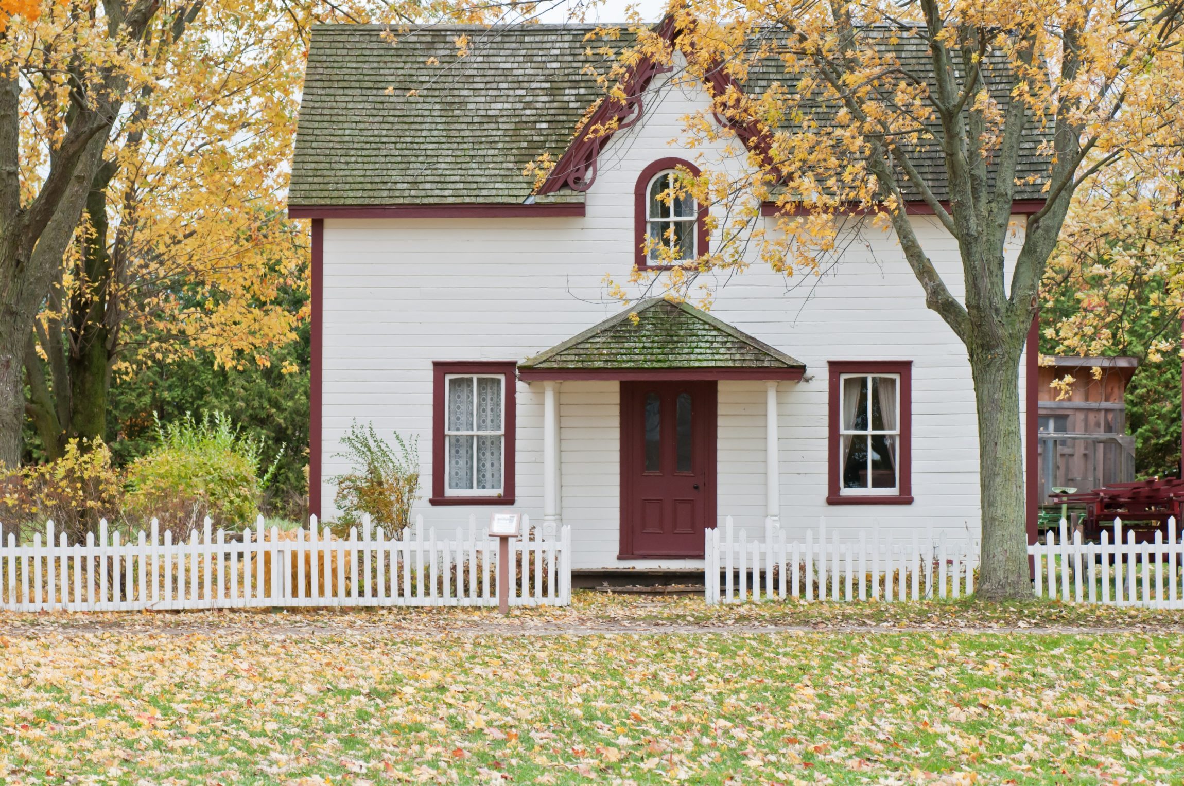 A white house and a white picket fence with trees and leaves on the grass.